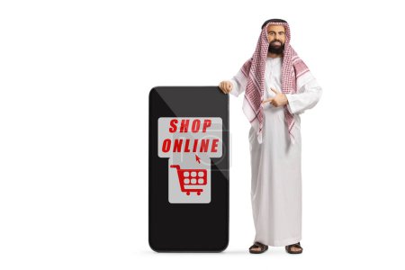 Foto de Hombre árabe saudí con ropa étnica de pie junto a un teléfono móvil con un cartel de compras en línea aislado sobre fondo blanco - Imagen libre de derechos