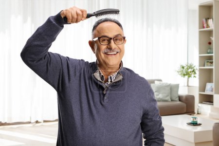 Photo for Mature man holding a hair brush bove head at home in a living room and smiling at camera - Royalty Free Image
