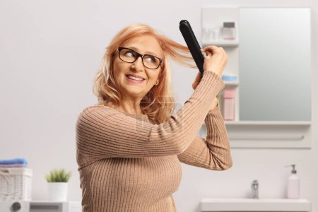 Photo for Middle aged woman using a wireless hair straightener in a bathroom - Royalty Free Image