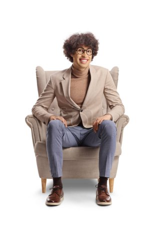 Foto de Joven con el pelo rizado y gafas sentado en un sillón y sonriendo aislado sobre fondo blanco - Imagen libre de derechos