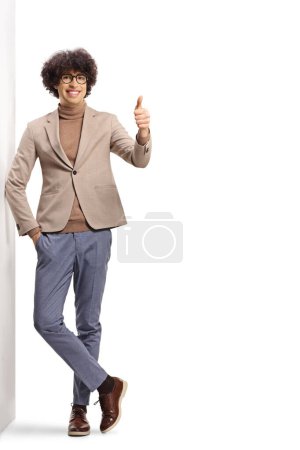 Photo for Full length portrait of a cheerful young man with glasses leaning on a wall and gesturing thumbs up isolated on white background - Royalty Free Image
