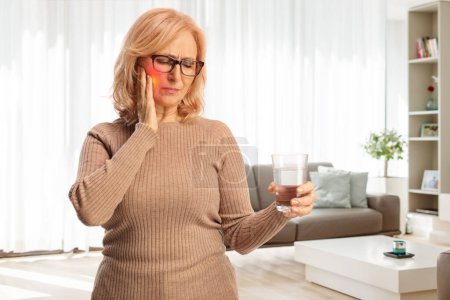 Téléchargez les photos : Femme d'âge moyen avec un mal de dents tenant un verre d'eau à la maison dans un salon - en image libre de droit