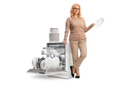Foto de Mujer de mediana edad sosteniendo un plato blanco limpio y de pie junto a un lavavajillas aislado sobre fondo blanco - Imagen libre de derechos