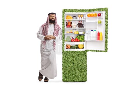 Photo for Full length portrait of a saudi arab man leaning on a power efficient fridge with food isolated on white background - Royalty Free Image