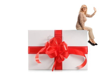 Foto de Mujer casual de mediana edad sentada en una gran caja de regalo y saludando aislada sobre fondo blanco - Imagen libre de derechos
