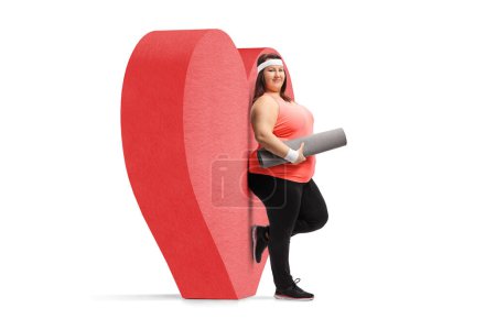 Foto de Mujer de talla grande con una esterilla de ejercicio apoyada en un corazón rojo aislado sobre fondo blanco - Imagen libre de derechos