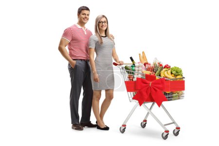 Foto de Joven hombre y mujer posando con un carrito de compras completo y atado con una cinta roja aislada sobre fondo blanco - Imagen libre de derechos