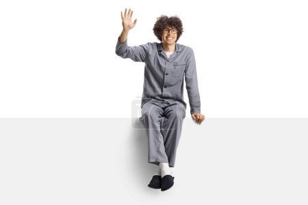 Photo for Young man in pajamas and slippers sitting on a blank panel and waving isolated on white background - Royalty Free Image