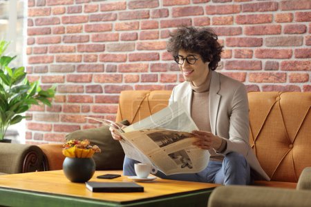 Téléchargez les photos : Jeune homme assis dans un café sur un canapé et lisant un journal - en image libre de droit