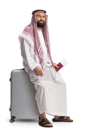 Téléchargez les photos : Saoudien arabe assis sur une valise et tenant un passeport isolé sur fond blanc - en image libre de droit