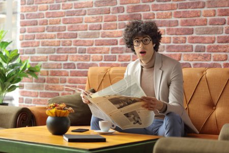 Foto de Sorprendido joven sentado en un café en un sofá y leyendo un periódico - Imagen libre de derechos