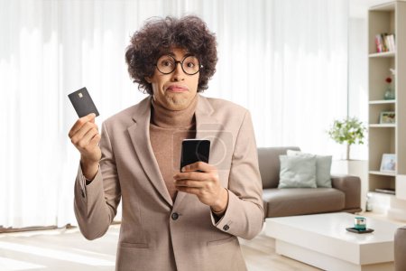 Téléchargez les photos : Jeune homme confus tenant une carte de crédit et un téléphone portable à la maison - en image libre de droit