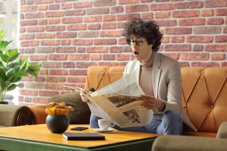 Téléchargez les photos : Jeune homme dans un café assis sur un canapé et lisant un journal - en image libre de droit
