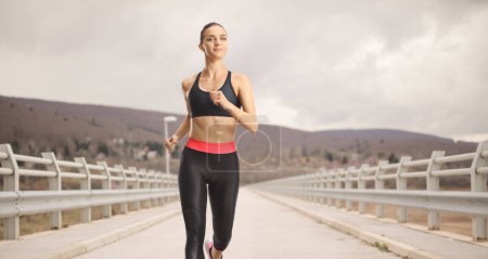 Téléchargez les photos : Jeune femme en forme courant sur un pont par une journée nuageuse - en image libre de droit