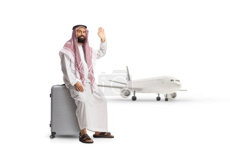Téléchargez les photos : Homme arabe saoudien assis sur une valise près d'un avion et agitant isolé sur fond blanc - en image libre de droit