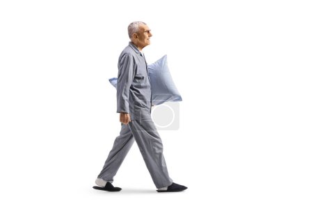 Téléchargez les photos : Homme âgé en pyjama portant un oreiller et marchant isolé sur fond blanc - en image libre de droit