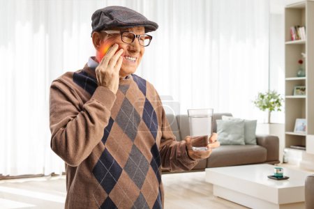 Téléchargez les photos : Homme âgé avec un mal de dents tenant un verre d'eau à la maison dans un salon - en image libre de droit