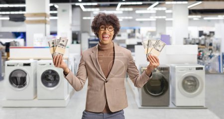 Photo for Young excited man holding stacks of money in a store with washing machines - Royalty Free Image