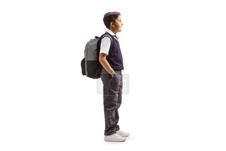 Full length profile shot of a schoolboy in a uniform standing with hands inside pockets isolated on white background