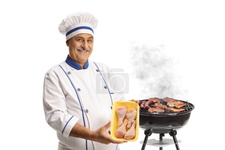Photo for Chef holding a pack of raw chicken meat next to a barbecue grill isolated on white background - Royalty Free Image
