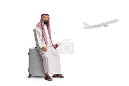 Téléchargez les photos : Saoudien arabe assis sur une valise avec passeport à la main et un avion décollant isolé sur fond blanc - en image libre de droit