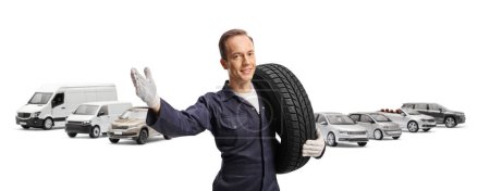 Foto de Mecánico de coche sosteniendo un neumático de coche y haciendo gestos con la mano delante de vehículos estacionados aislados sobre fondo blanco - Imagen libre de derechos