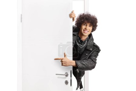Foto de Hombre joven en una chaqueta de cuero negro de pie detrás de una puerta y señalando - Imagen libre de derechos