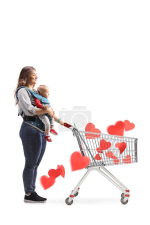 Photo for Full length shot of a mother with a baby in a carrier pushing a shopping cart full of red hearts isolated on white background - Royalty Free Image