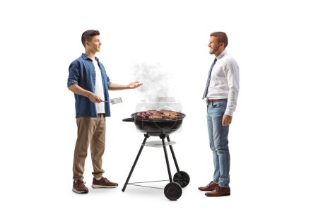 Téléchargez les photos : Jeune homme préparant de la viande sur un barbecue et parlant à un ami isolé sur fond blanc - en image libre de droit