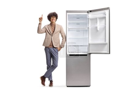 Téléchargez les photos : Jeune homme pointant vers le haut à côté d'un nouveau réfrigérateur isolé sur fond blanc - en image libre de droit