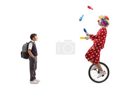Téléchargez les photos : Plan complet d'un écolier surpris regardant un clown jongler et chevauchant un monocycle isolé sur fond blanc - en image libre de droit