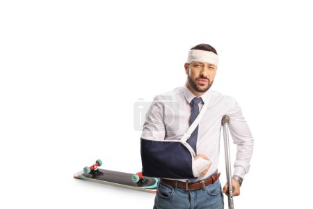Téléchargez les photos : Homme avec une fronde de bras et bandage sur la tête appuyé sur une béquille, concept de blessure de planche à roulettes isolé sur fond blanc - en image libre de droit
