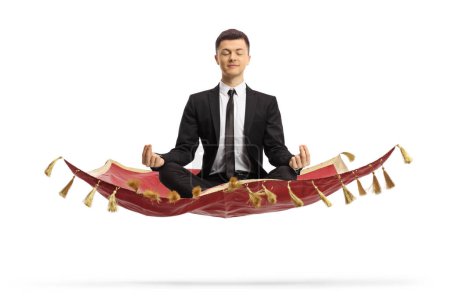 Photo for Young man in a black suit sitting on a flying carpet and meditating isolated on white background - Royalty Free Image