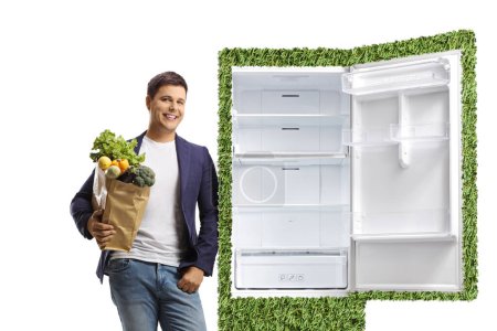Jeune homme tenant un sac d'épicerie et s'appuyant sur un réfrigérateur écologique vert isolé sur fond blanc