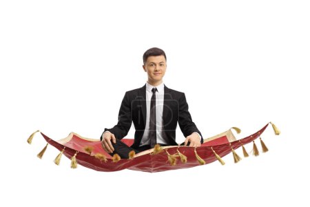 Téléchargez les photos : Young man in a black suit sitting with crossed legs and meditating on a magic carpet isolated on white background - en image libre de droit