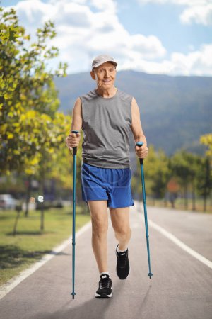 Téléchargez les photos : Homme âgé en vêtements de sport marchant avec des bâtons de trekking dans un parc - en image libre de droit