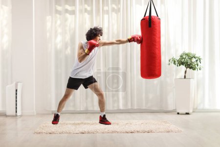 Téléchargez les photos : Jeune homme frappant un sac à la maison avec des gants de boxe - en image libre de droit