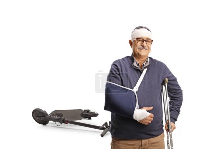 Téléchargez les photos : Homme mûr avec un bras cassé et bandage sur la tête appuyé sur une béquille, concept de blessure scooter électrique isolé sur fond blanc - en image libre de droit