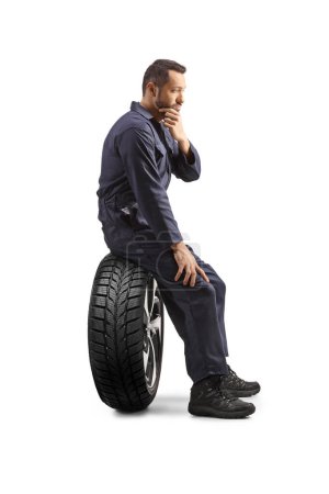 Téléchargez les photos : Mécanicien automobile pensif en uniforme assis sur un pneu isolé sur fond blanc - en image libre de droit