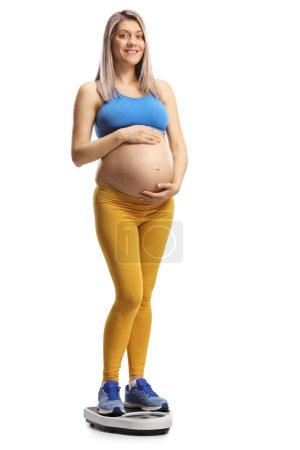 Téléchargez les photos : Femme enceinte en tenue de sport tenant son ventre et debout sur une échelle de poids isolée sur fond blanc - en image libre de droit