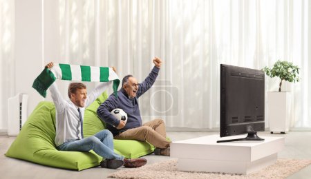 Téléchargez les photos : Des hommes à la maison applaudissant avec une écharpe et un ballon, assis sur un fauteuil de sac de haricot et regardant le football - en image libre de droit