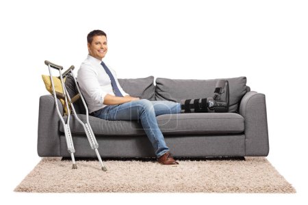 Téléchargez les photos : Homme d'affaires avec une blessure au pied et attelle de jambe assis sur un canapé isolé sur fond blanc - en image libre de droit