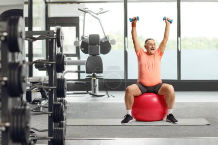 Téléchargez les photos : Homme mûr souriant en tenue de sport assis sur une balle de fitness et faisant de l'exercice dans une salle de gym - en image libre de droit