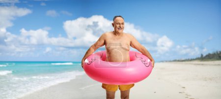 Foto de Hombre maduro sonriente que usa pantalones cortos de natación y anillo de goma inflable de flamenco en una playa de arena - Imagen libre de derechos