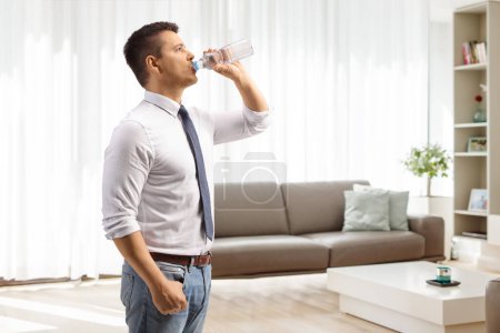 Téléchargez les photos : Homme buvant l'eau d'une bouteille en plastique à la maison dans un salon - en image libre de droit