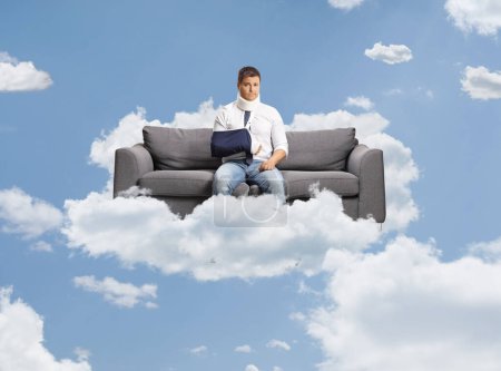 Trauriger Mann mit gebrochenem Arm und Halswirbel, der auf einem Sofa sitzt, das im Himmel schwebt