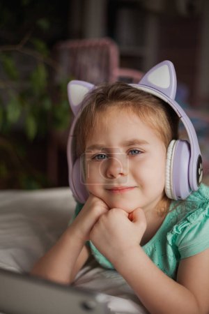 Photo for Adorable little girl of 4 years old is watching video on tablet with wireless children's headphones. Video communication. Listening to music. - Royalty Free Image