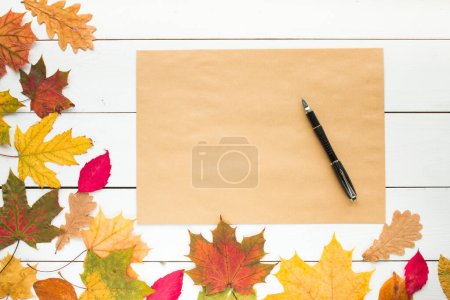 Téléchargez les photos : Feuille de papier artisanal et stylo avec des feuilles tombées sur fond blanc en bois, vue de dessus, espace de copie. espace libre pour le texte - en image libre de droit