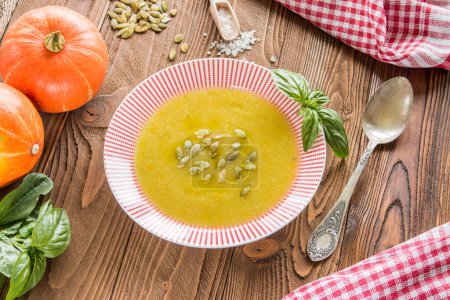 Photo for Cream soup from pumpkin and other vegetables decorated by basil leaf and pumpkin seeds, basil, pumpkins fruits, salt, pumpkins  seeds  on wooden table with silver spoon on red squared  napkin - Royalty Free Image