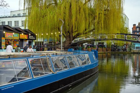 Photo for Camden Town, London, UK: A boat on Regents Canal near Camden Market. With a willow tree, footbridge, people and food outlets. - Royalty Free Image